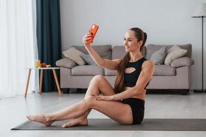 With phone in hands. Young woman with slim body type and in yoga clothes is at home photo