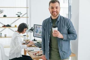 hombre positivo parado frente a una mujer sentada junto a la mesa. dos diseñadores trabajando juntos en la oficina foto