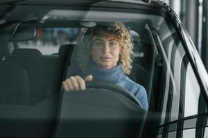 Sitting at driver's seat. Woman with curly blonde hair is in autosalon photo