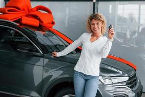 Car is with gift length on the top. Woman with curly blonde hair is in autosalon photo