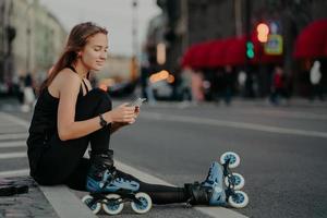 una foto al aire libre de una joven deportiva activa vestida con ropa activa usa patines revisa el contenido del mensaje en poses de teléfonos inteligentes contra el fondo borroso de la ciudad en el asfalto lleva un estilo de vida saludable