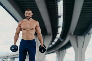 Serious bearded European man with sporty body prepares for barbell workout stands under bridge outdoor has strong biceps and arms concentrated down. Bodybuilding power lifting training concept photo