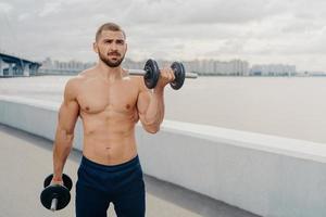 Handsome bearded man pumps up muscles, does exercises with barbells, leads sporty lifestyle, has muscular strong body, stands outdoor near river. Bare chested bodybuilder lifts heavy dumbbells photo