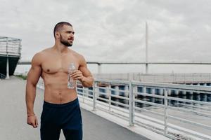 Handsome bearded sportsman drinks water from bottle promots healthy lifestyle and concentrated away into distance, relaxes after morning run, poses outside near river, being thirsty and tired. photo