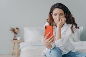 Stressed caucasian woman is doom scrolling. Girl reading morning news on smartphone in her bedroom. photo
