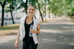 Outdoor shot of glad sporty woman drinks fresh water from bottle has walk during summer green urban park has toothy smile on face leads healthy lifestyle. Restoring aqua balanace after training