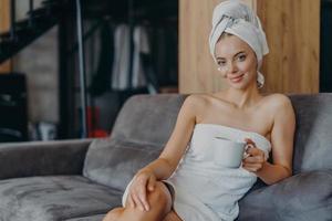 Relaxed beautiful young woman has healthy skin wears soft towel on head and around naked body holds mug of coffee poses at sofa with aromatic beverage enjoys weekends has well groomed complexion photo