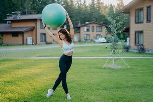 Slim young European woman holds fitball over head, dressed in cropped top and leggings, has gymnastics exercises outdoor, poses at green lawn near private house. People, sport, training concept photo