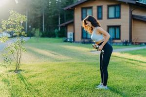 Full length shot of active sporty young woman raises dumbbells has workout outdoor near house stands on green lawn, dressed in sportsclothes, tries to be in good shape, stays fit and healthy photo