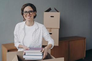 Happy nerd in glasses is stacking books into boxes. Young spanish woman packing carton container. photo