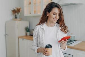 European woman with smartphone has coffee. Girl is reading message in kitchen at home and smiling. photo