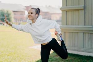 Outdoor shot of pretty young Eruopean woman in sportswear does warm up exercises outdoors stretches legs before run looks into distance with happy expression listens music in wireless earphones photo