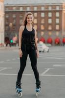Positive young female model in sportsclothes rides on blades enjoys leisure activities poses at urban place against blurred background stands in full length. Active lifestyle and rollerblading concept photo
