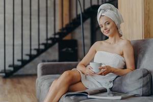 Horizontal shot of pleased young European woman smiles gently poses wrapped in towel drinks coffee poses on couch in living room reads magazine, applies beauty cream on face after taking shower photo