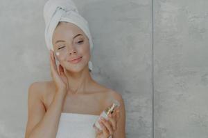Lovely European woman applies facial moisturizer holds bottle of body lotion, has healthy skin, well groomed complexion wears wrapped towel on head after taking shower, poses against grey background photo