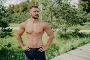 Horizontal shot of thoughtful shirtless sportsman keeps hands on waist, looks away demonstrates his muscular body, poses over nature background. Active bodybuilder goes in for sport, has daily workout photo