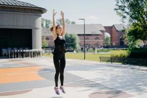 Positive active female model has cardio training outdoors jumps high keeps arms raised dressed in activewear being strong fit spends free time on stadium has regular morning workout. Fitness and sport photo