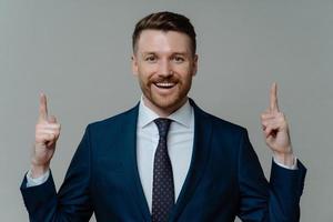 Pleased businessman in suit pointing up with fingers at copy space photo