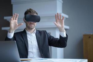 Man CEO in virtual reality glasses with hands up in air at home office interior photo