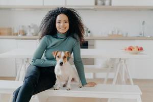Happy housewife with Afro haircut, sits at bench with pedigree dog, have fun and look directly at camera, pose in kitchen, express good emotions, relax together. Lovely pet with owner at home photo