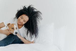 Indoor shot of lovely Afro American girl rests in bed after awakening with dog, enjoys time with pet, sit on comfortable bed against white wall. Jack russell terrier pays with owner. Cute friendship photo