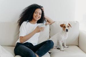 Beautiful woman with Afro haircut, drinks coffee, poses in living room at sofa with pedigree dog, wears white t shirt and jeans, enjoys coziness and comfort at home. People, animals, lifestyle photo