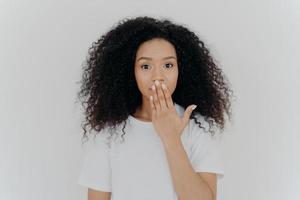 Headshot of impressed curly young woman covers mouth with palm, tries to be speechless, has curly hairstyle, manicure and minimal makeup, wears white t shirt, poses indoor. Omg, what gossip photo