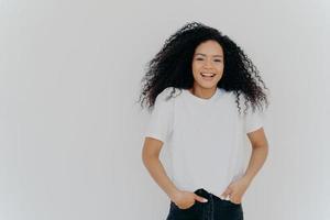 Half length shot of good looking smiling woman laughs at funny joke, has fun, keeps both hands in pockets of jeans, wears white t shirt, has curly fluffy hair, poses indoor, blank space on right side photo
