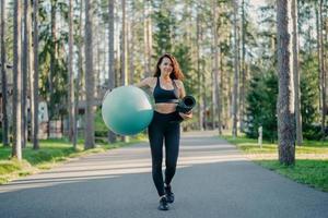 Horizontal shot of active slim woman dressed in sportswear, carries fitball and rolled up karemat, walks on road near trees, going to have workout, breathes fresh air, leads healthy sporty lifestyle photo