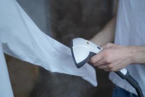 Cropped photo of man using steamer neatly ironing white shirt while holding long sleeve