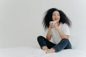 Lazy morning and bedtime concept. Positive Afro woman holds mug of hot tea or coffee, sits in bed, looks gladfully aside, enjoys good weather, wears comfortable clothes, white wall in background photo