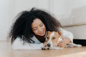 Relaxed Afro woman with crisp dark hair lies on floor, plays with cute puppy, has fun with jack russell terrier dog wears white sweater being in living room. Happy owner petting lovely domestic animal photo