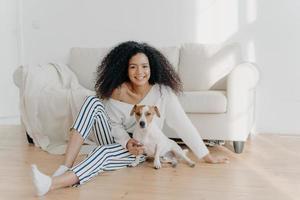 una joven afroamericana relajada se sienta en el piso en una habitación vacía cerca del sofá con un perro pedigrí, disfruta de un momento dulce en casa, se muda a un nuevo apartamento para vivir. dama descansa con cachorro en la sala de estar foto