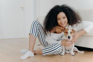 Indoor shot of pleased curly haired female model poses for photo with jack russell terrier dog, embraces pet tenderly, sits on floor with beloved animal near comfortable sofa, spend time together