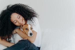 Photo of attractive woman with curly Afro hairstyle, cuddles and pets dog with smile, expresses love, enjoys cozy domestic atmosphere, pose against white background with empty space for promotion