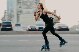 Young dark haired slim healthy woman has recration time enjoys rolleblading on speed moves fast has cheerful expression wears black t shirt and leggings. Active rest and sport training concept photo