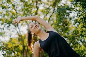 Sporty fitness woman does warm up and stretching exercises leans aside has content face expression dressed in active wear poses against nature background during summer day. Sport and motivation photo