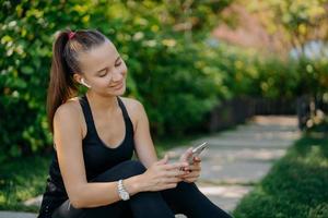 Pleased active woman with pony tail wears sportive wear concentrated at smartphone device listens music in wireless earphones browses internet has outdoor training. Healthy lifestyle concept photo