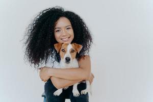 People, animal or pet care concept. Curly haired woman embraces favourite dog, smiles pleasantly, stands against white background, copy space area for your advertising content. Good friends. photo