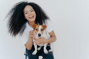 Portrait of joyful curly girl petting her dog, rejoicing buying jack russell terrier, smiles broadly, plays with animal, wears casual clothing, isolated over white background, enjoys good day photo