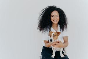 concepto de humanos y animales. una mujer alegre y guapa con el pelo terso, sonríe agradablemente, juega con un perro pedigrí, se sienta en una silla cómoda, hace una toma memorable, posa contra un fondo blanco foto