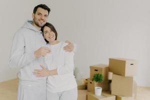 Couple in love purchase own house, embrace and stand closely to each other, hold keys from new apartment, carton boxes in background, have delighted face expressions. Satisfied clients move in photo