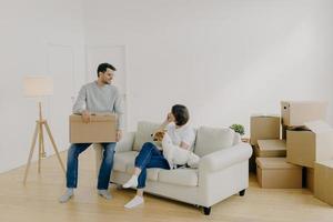 Positive woman and man pose in empty spacious room during relocation day, husband carries cardboard boxes with belongings, wife has telephone conversation, sits on comfortable sofa with dog. photo