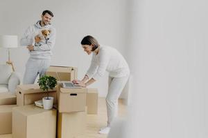 Indoor shot of attractive woman leans to laptop computer, focused into screen, surfes internet, searches ideas for decoration, man stands near, plays with favourite pet, move into new house. photo