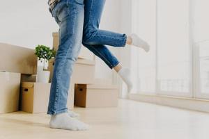 Happy unknown couple move in new abode, man lifts woman, wear jeans, pose in empty room with carton boxes around, start living seperate from parents rejoice buying apartment. Unrecognizable homeowners photo