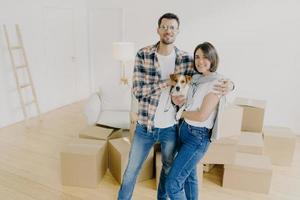 felices inquilinos de mujeres y hombres o inquilinos de piso posan en su propia casa, abrazan y posan con un perro pequeño, tienen expresiones alegres, comienzan a vivir en un apartamento recién comprado. concepto de familia y reubicación. foto