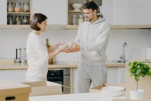 Horizontal shot of smiling couple unpack cardboard boxes in new kitchen, man passes plates to woman, placing dishes in cupboard, surrounded with moving packages in own bought or rented apartment photo