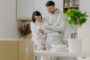 Horizontal view of happy young woman and man play with dog, drink takeaway coffee, stand against kitchen interior, unpack boxes with personal stuff, wear casual clothes, bought new apartment photo