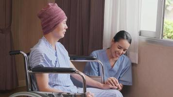 Uniformed young Asian female therapy doctor holding hands wheelchair male patient at window to support and motivate recovery, cancer illness after chemo medical treatment in hospital inpatient room. video