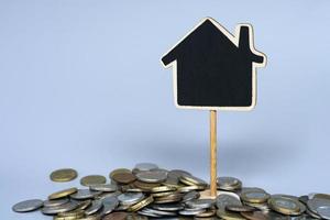 Wooden house model with pile of coins against blue background. Copy space. photo